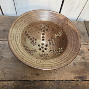 May include: A round, speckled brown ceramic berry bowl featuring a wide, ridged rim and a series of small, circular drainage holes arranged in a floral pattern across the interior base and sides, showcasing a natural, earthy glaze finish.