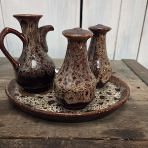 May include: A ceramic serving set featuring a pitcher and two matching bottles, all with a speckled brown glaze. The set rests on a round tray with a similar speckled pattern. The pieces are displayed on a rustic wooden surface.