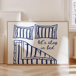 Puede incluir: Una impresión artística enmarcada que presenta una cama con almohadas y ropa de cama con un diseño de rayas azul marino sobre un fondo blanco. El texto "let's stay in bed" está escrito en cursiva. La obra de arte se exhibe en una habitación con luz natural.