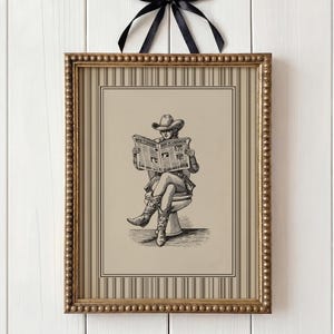 May include: A framed sepia-toned print depicting a person in cowboy attire reading a newspaper whilst seated on a toilet. The gold frame has a black bow at the top. The print is hung on a white wall.