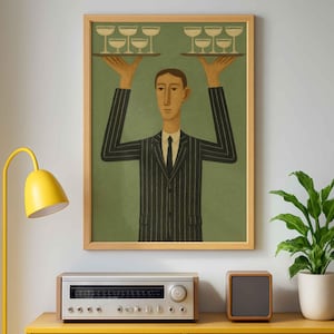 Puede incluir: Una impresión artística enmarcada representa a una persona con un traje de rayas llevando bandejas de copas de champán. La obra de arte tiene un fondo verde y un marco de madera clara. La impresión está encima de un mueble amarillo con un equipo de música y un altavoz pequeño.