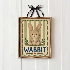May include: A framed print with a cartoon rabbit and the word "WABBIT". The rabbit is brown with pink ears, set against a green and cream striped backdrop. A black ribbon is attached to the top. The frame has a gold-colored beaded edge.