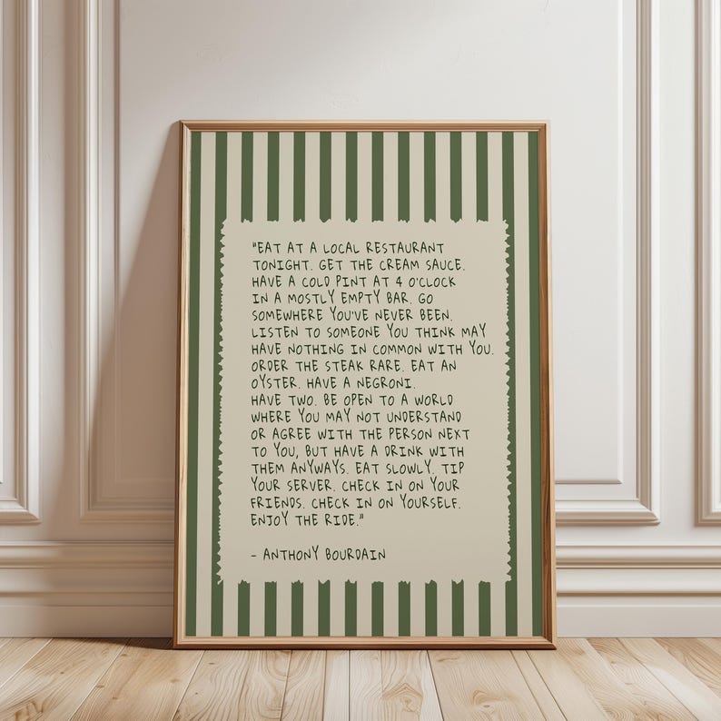 May include: A framed print with a wooden frame. The print features a cream-colored background with text from Anthony Bourdain, surrounded by a green and white striped border. The text is in black and is a quote about life and experiences.