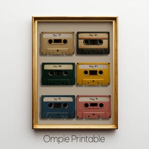 Puede incluir: Una impresión enmarcada con seis cintas de cassette vintage en varios colores: beige, negro, verde, amarillo, azul y rosa. Cada cinta tiene una fecha escrita a mano. El marco es dorado y la impresión está etiquetada "Ompie Printable" en la parte inferior.