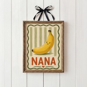 May include: A framed print with a smiling yellow banana illustration. The artwork has a green and beige striped background, with the word "NANA" in orange letters at the bottom. A black ribbon is tied at the top.