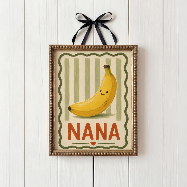 May include: A framed print with a smiling yellow banana illustration. The artwork has a green and beige striped background, with the word "NANA" in orange letters at the bottom. A black ribbon is tied at the top.