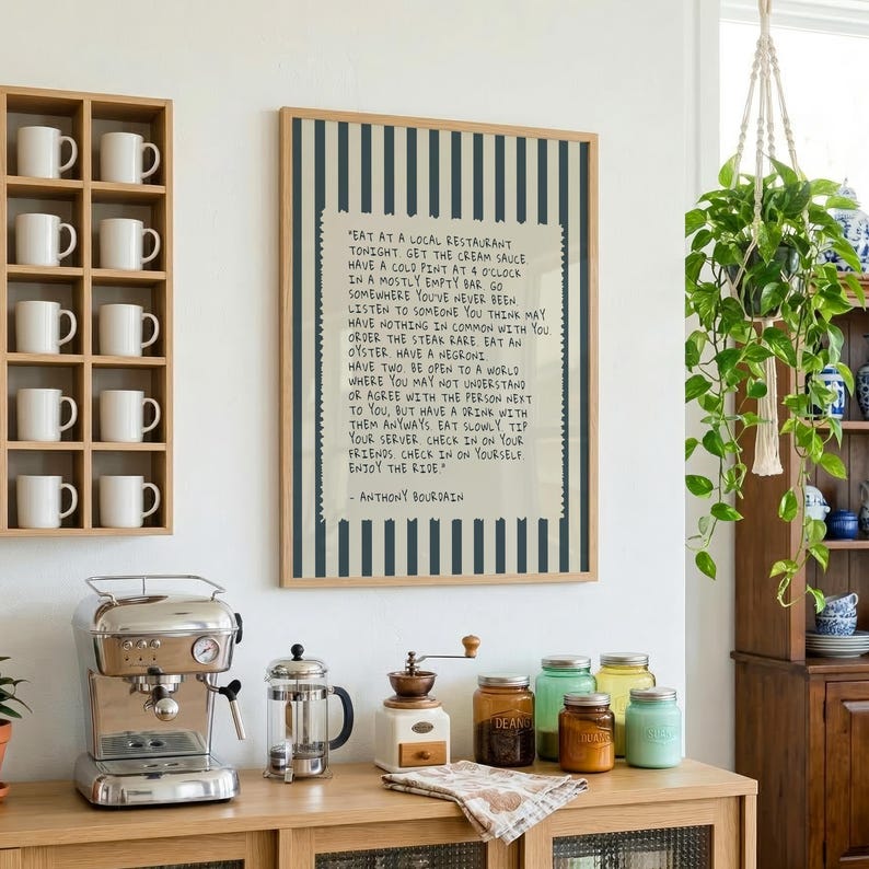 Striped Anthony Bourdain typography poster with handwritten text on a warm cream background featuring olive green, navy blue, and charcoal black colour variations. Vintage-inspired kitchen wall art with subtle stripe details and European cafe aesthetic displayed in wooden frames inside cozy interiors. Minimal quote print designed for coffee corners, dining rooms, bookstores, restaurants, and modern apartments with warm lighting and neutral decor styling.