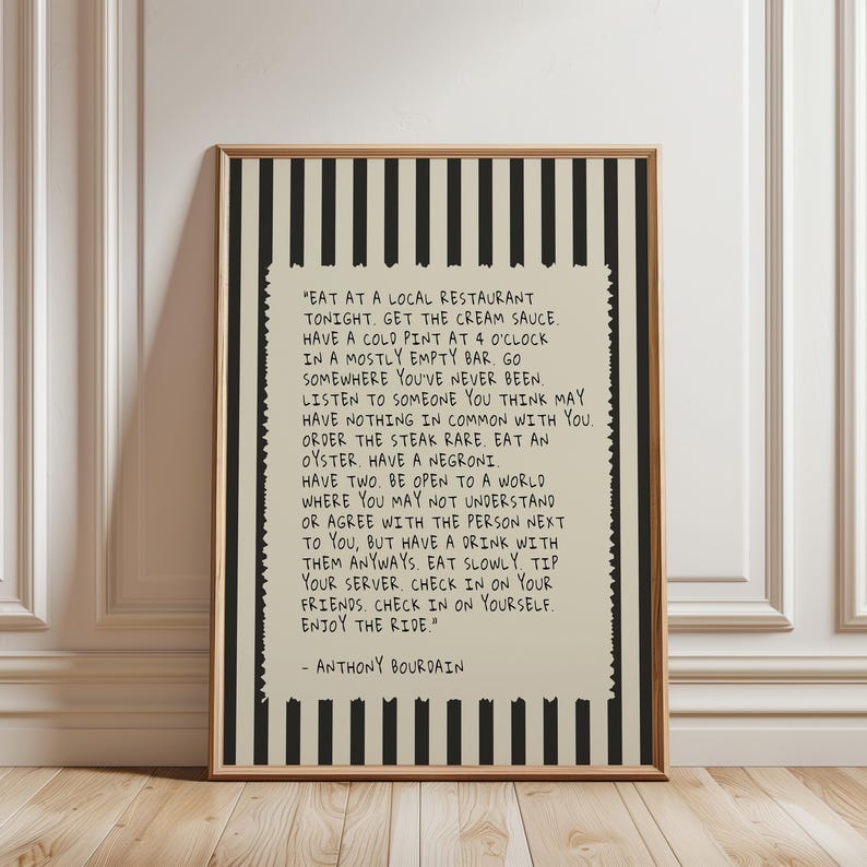 May include: A framed print with a beige background and black vertical stripes. The print features a quote by Anthony Bourdain in black text. The quote is surrounded by a decorative border and framed in a wooden frame.