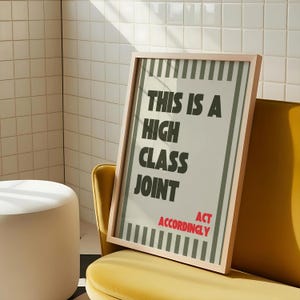 May include: A framed poster with the text "THIS IS A HIGH CLASS JOINT" in black, and "ACT ACCORDINGLY" in red. The poster has a light beige background with vertical green stripes and is in a light wood frame.