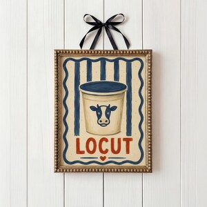 May include: A framed print with a vintage illustration of a dairy product container. The container is beige with a blue lid and a cow graphic. The word "LOCUT" is in red, with a heart below. A black ribbon is tied at the top.