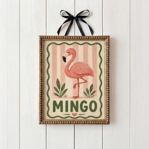 May include: A framed print with a pink flamingo illustration on a pink and white striped background. The word "MINGO" is written in green below the flamingo, with a heart symbol. The frame is gold, and a black ribbon is attached.