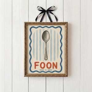 May include: A framed print with a spoon illustration. The artwork has a gold-coloured frame and a blue and white striped background. The word "FOON" is written in red letters at the bottom. A black ribbon is tied at the top.