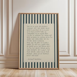 May include: A framed print with a beige background and dark blue vertical stripes. The print features a quote by Anthony Bourdain in black text, with a decorative border. The quote encourages trying new experiences and enjoying life.