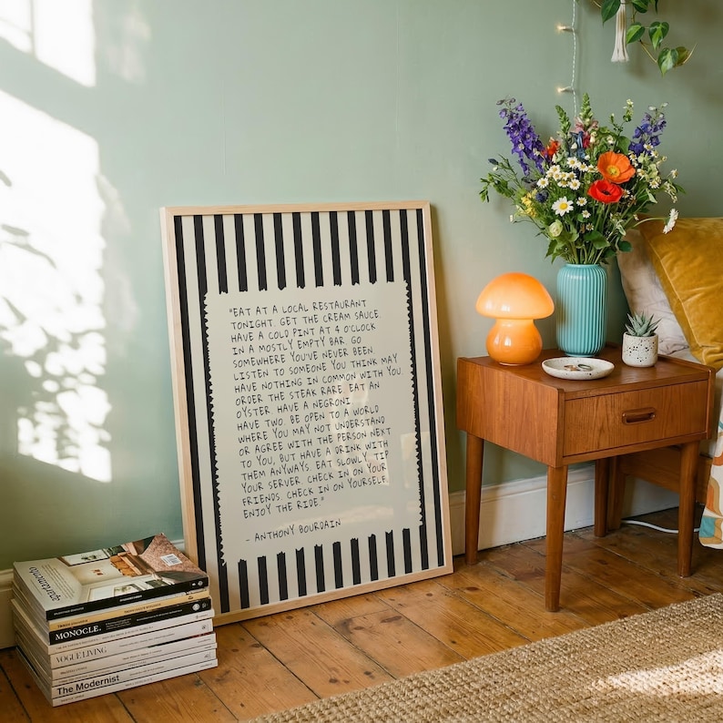 Striped Anthony Bourdain typography poster with handwritten text on a warm cream background featuring olive green, navy blue, and charcoal black colour variations. Vintage-inspired kitchen wall art with subtle stripe details and European cafe aesthetic displayed in wooden frames inside cozy interiors. Minimal quote print designed for coffee corners, dining rooms, bookstores, restaurants, and modern apartments with warm lighting and neutral decor styling.