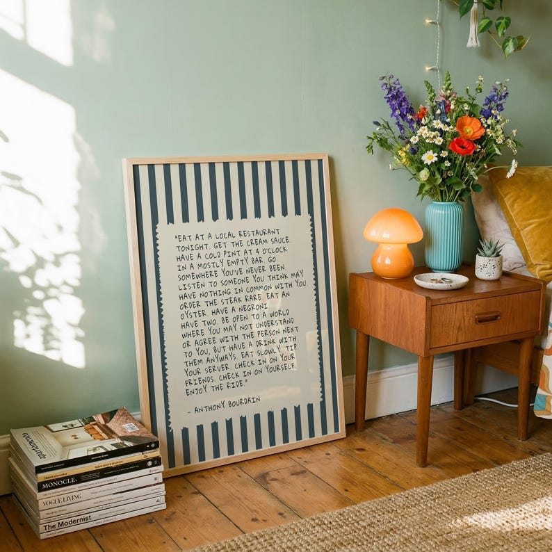 Striped Anthony Bourdain typography poster with handwritten text on a warm cream background featuring olive green, navy blue, and charcoal black colour variations. Vintage-inspired kitchen wall art with subtle stripe details and European cafe aesthetic displayed in wooden frames inside cozy interiors. Minimal quote print designed for coffee corners, dining rooms, bookstores, restaurants, and modern apartments with warm lighting and neutral decor styling.