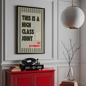 May include: A framed poster with the text "THIS IS A HIGH CLASS JOINT ACT ACCORDINGLY" hangs above a red cabinet. A black record player sits on the cabinet. A white globe light and a glass vase with branches are also in the image.