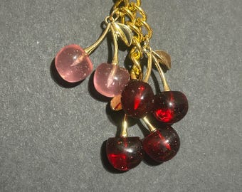 May include: A gold-coloured keyring featuring a cluster of cherries. The cherries are a mix of translucent pink and deep red, with gold-coloured stems and leaves. The keyring has a gold ring and chain.
