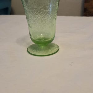 May include: A light green translucent glass tumbler featuring a pedestal base and an intricate embossed floral pattern covering the exterior surface. The glass has a flared rim and a circular foot, showcasing a detailed relief design throughout.
