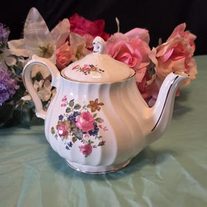May include: A white porcelain teapot with a ribbed body and a floral pattern featuring pink roses, blue flowers, and green leaves. The teapot has a matching lid with a small floral design and gold trim on the handle, spout, lid, and base edges.