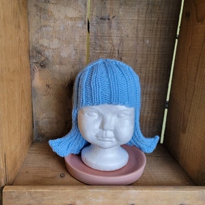 May include: A light blue knitted hat with attached hair, displayed on a white mannequin head. The hat is ribbed and sits on a small, pink, round dish. The background is a wooden box.