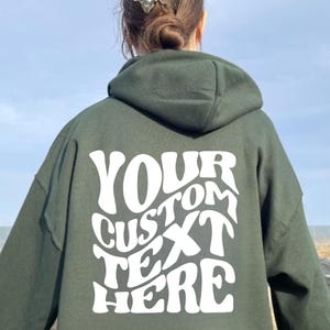 May include: Dark green hoodie with the text "YOUR CUSTOM TEXT HERE" in white, displayed on the back. The hoodie has a hood and long sleeves. The image is taken outdoors.