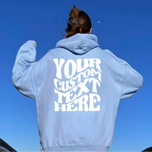 May include: A light blue hoodie with the text "YOUR CUSTOM TEXT HERE" in white, displayed on the back. The hoodie has a hood and long sleeves. The background is a clear, bright blue sky.