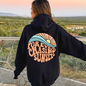May include: Black hoodie with a colorful graphic on the back. The graphic features a stylized sunset with the words "Forever Chasing Sunsets" in a retro font. The hoodie has a hood and long sleeves.