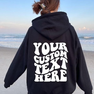 May include: Black hoodie with the text "YOUR CUSTOM TEXT HERE" in white, displayed on the back. The hoodie is worn by a person standing on a beach with the ocean in the background. The hoodie has a hood and long sleeves.