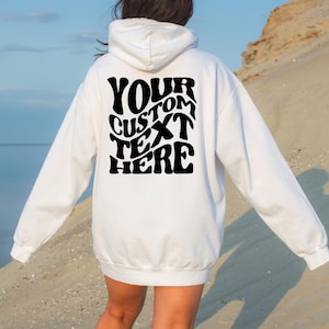 May include: White hoodie with a hood and long sleeves. The back of the hoodie features the text "YOUR CUSTOM TEXT HERE" in a black, retro-style font. The hoodie is made of a soft material and is suitable for casual wear.