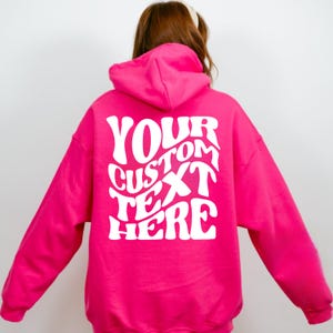 May include: A bright pink hoodie with the text "YOUR CUSTOM TEXT HERE" in white, retro-style lettering on the back. The hoodie has a hood and long sleeves. The image is taken from the back.