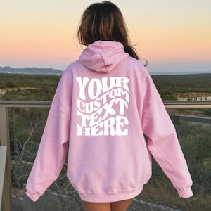 May include: A light pink hoodie with a drawstring hood and long sleeves. The back of the hoodie features the white text "YOUR CUSTOM TEXT HERE" in a wavy, retro font. The background shows a scenic view of a landscape.