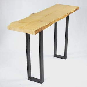 May include: A light-colored wooden console table with a live edge top. The table has a rectangular shape and is supported by black metal legs. The natural wood grain is visible on the tabletop. The table is approximately 60 inches long.