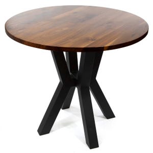 May include: A round table featuring a smooth, circular wooden top with a visible natural grain pattern in a medium brown tone. The table is supported by a central base consisting of four angled, matte black metal legs that intersect at the center.