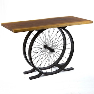 May include: A unique console table with a rectangular wooden top and a base made from two repurposed bicycle wheels. The wheels are black and support the brown wooden tabletop. The table is a stylish piece of furniture.