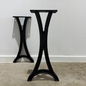 May include: Two black cast iron table legs. Each leg has a distinctive V-shape design, with a flat top and a curved base. The legs are designed for stability and support, suitable for various table sizes.