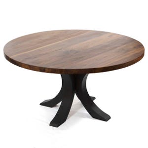 May include: A round wooden dining table with a dark brown tabletop and a black pedestal base. The tabletop appears to be made of solid wood, with visible grain patterns. The base has a unique, curved design. The table is suitable for a dining room or kitchen.