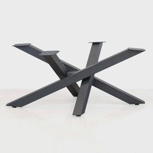 May include: A dark gray metal table base with a modern, geometric design. The base features four angled legs that intersect in the center, creating a starburst pattern. The top has four mounting plates for attaching a tabletop. The base is suitable for a dining or coffee table.