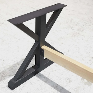 May include: Black metal table base with an X-shaped design. The base has a rectangular top with pre-drilled holes for mounting. A light-colored wooden beam is attached to the base. The base is designed to support a table top.