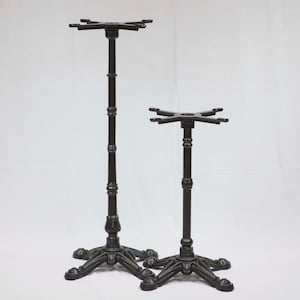 May include: Two black cast iron table bases. One base is taller than the other. Each base has a decorative, ornate footed base and a top with four arms to support a tabletop. The bases are made of metal.