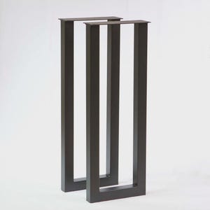 May include: Two dark gray metal table legs with a rectangular design. Each leg has a flat top and a square base, with three vertical supports. The legs are designed for a modern aesthetic.