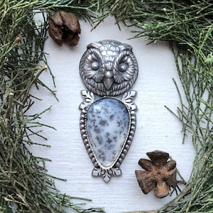 May include: A silver owl pendant featuring a white and black agate stone in the centre. The pendant is surrounded by green pine branches.