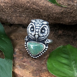 Op de afbeelding: Een zilveren ring met een uil en een groene edelsteen. De ring ligt op een bruine steen met groene bladeren.