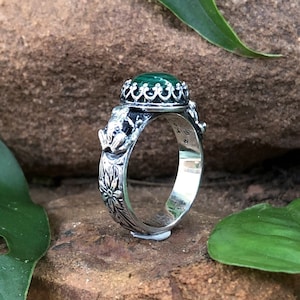 Puede incluir: Un anillo de plata con una piedra preciosa verde engastada en un diseño de corona. La banda está decorada con motivos florales.