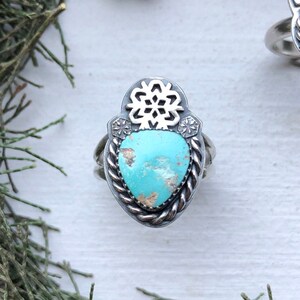 Pode incluir: Duas alianças de prata com pedras turquesa engastadas em um design de floco de neve. As alianças estão sobre um fundo branco com galhos de pinheiro verdes.