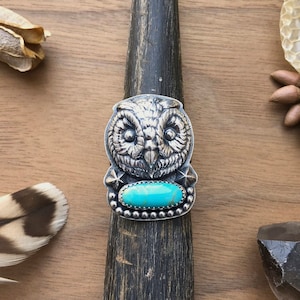 May include: A silver ring with an owl design and a turquoise stone. The ring is on a wooden surface.