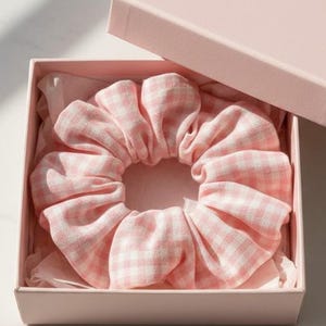 Puede incluir: Una goma para el pelo rosa a cuadros vichy dentro de una caja de regalo rosa a juego. La goma para el pelo es de tela suave con un estampado de cuadros blancos y rosas. La caja está abierta.