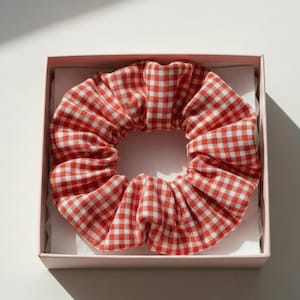 Könnte beinhalten: Ein rot-weiß kariertes Haargummi liegt in einer hellrosa Schachtel. Das Haargummi aus Stoff ist gerafft und bildet eine volle, kreisförmige Form. Die quadratische Schachtel hat ein schlichtes Design.
