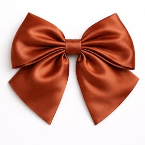 May include: A large, satin bow in a rich, warm brown colour. The bow is symmetrical, with two large loops and two long tails. The fabric has a slight sheen, and the bow is likely used for decoration or gift wrapping.