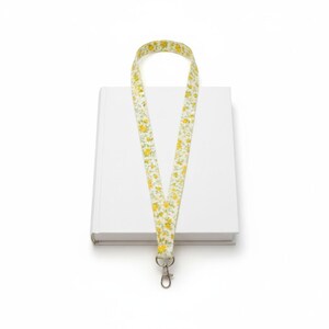 Pode incluir: Um livro branco com um cordão floral. O cordão tem um fundo creme com flores amarelas e folhas verdes. Possui um fecho prateado.