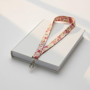Pode incluir: Um cordão floral com um fecho de metal repousa sobre um livro branco fechado. O cordão apresenta um fundo creme com rosas rosa e vermelhas e folhas verdes. O livro tem uma capa branca lisa e uma lombada cinza.